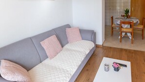 A modern living room with a grey sofa, two pink cushions, a white quilted mattress, a wooden coffee table with a remote control, a vase with flowers, and a dining area with a wooden table and chairs.