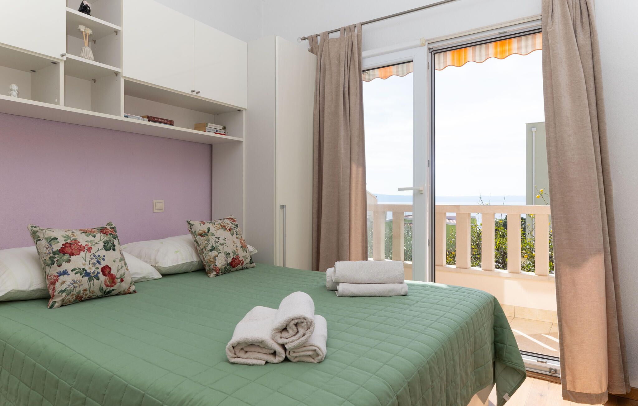 A bedroom with a green bedspread, floral pillows, a white wardrobe, and a balcony with curtains.