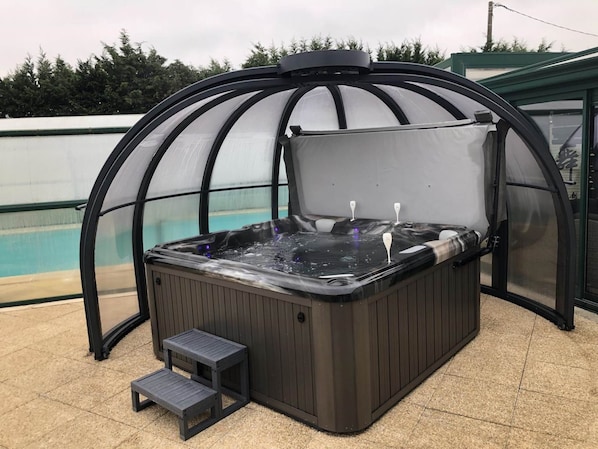 A hot tub with a glass enclosure, two steps leading up to it, and a pool in the background.