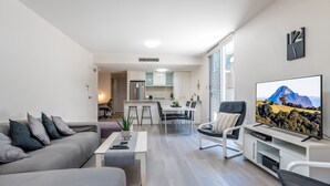 A modern living room with a sofa, coffee table, and TV stand. There is a kitchen in the background with a dining area. - City Edge Albion Pool, Patio & City Access (Albion)
