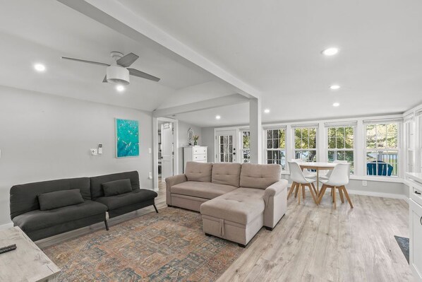 Un vaste espace de vie comprenant un grand canapé d’angle, une table basse et une table à manger avec des chaises. On y trouve également un ventilateur de plafond, un tapis et une vue sur l’extérieur grâce à de grandes fenêtres.