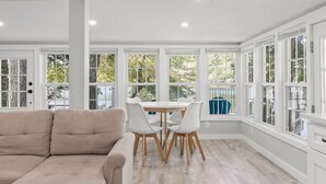 A bright room with a white couch, a round table with chairs, and large windows with white frames.