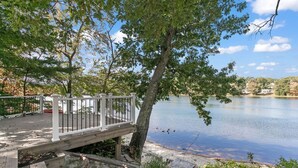 A wooden deck with a white railing overlooking a calm lake, surrounded by trees and a clear sky.