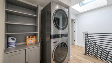 Un espace buanderie comprenant une machine à laver et un sèche-linge superposés, une étagère avec des produits de nettoyage et une lucarne au-dessus.
