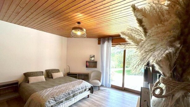A bedroom with a wooden ceiling, a bed with a quilted cover, a chair, and a large window with a view of greenery.