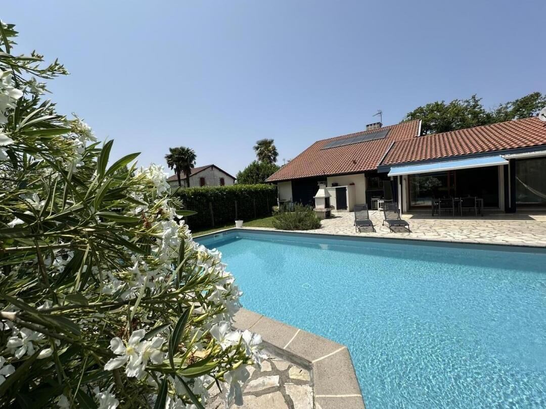 A swimming pool with a tiled deck, a covered outdoor seating area, and a house with a red-tiled roof in the background.