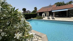 A swimming pool with a tiled deck, a covered outdoor seating area, and a house with a red-tiled roof in the background.