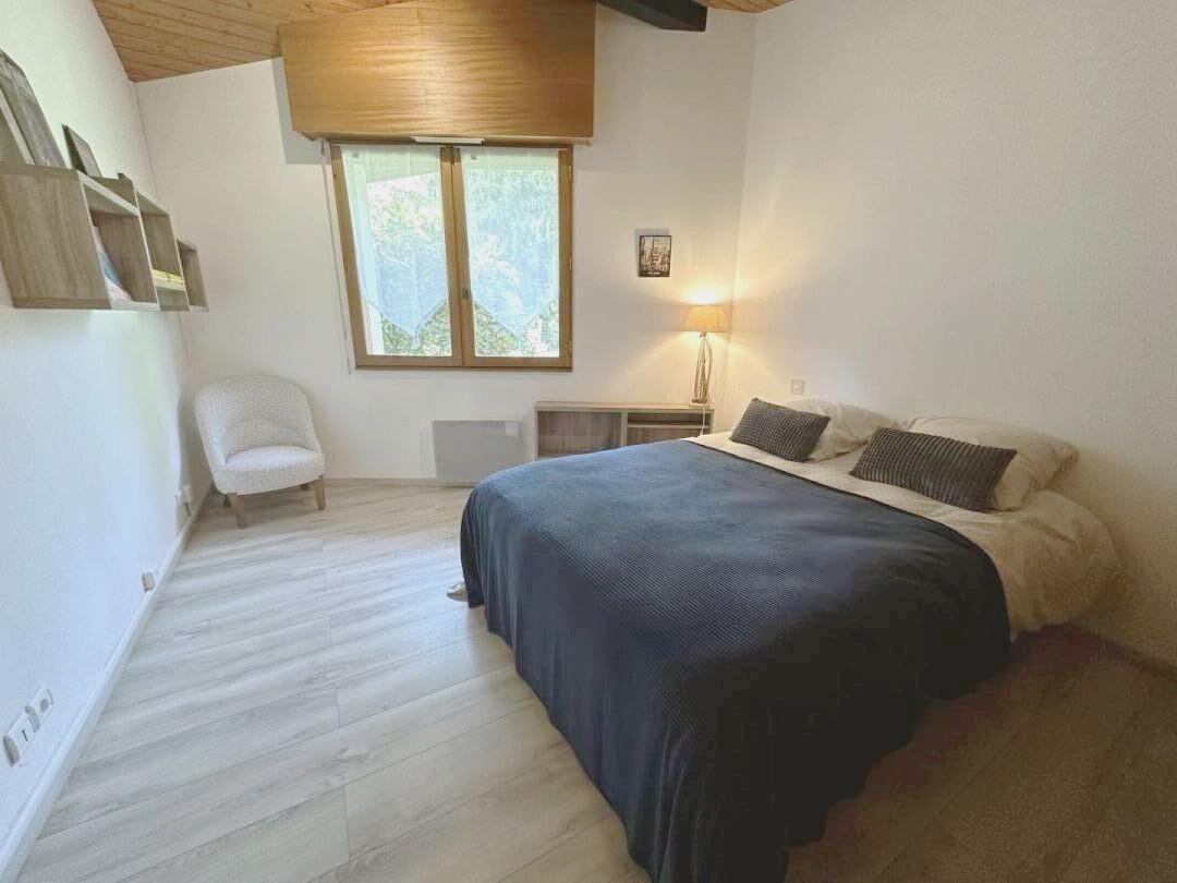 A bedroom with a bed, a chair, a bedside table with a lamp, and a window.