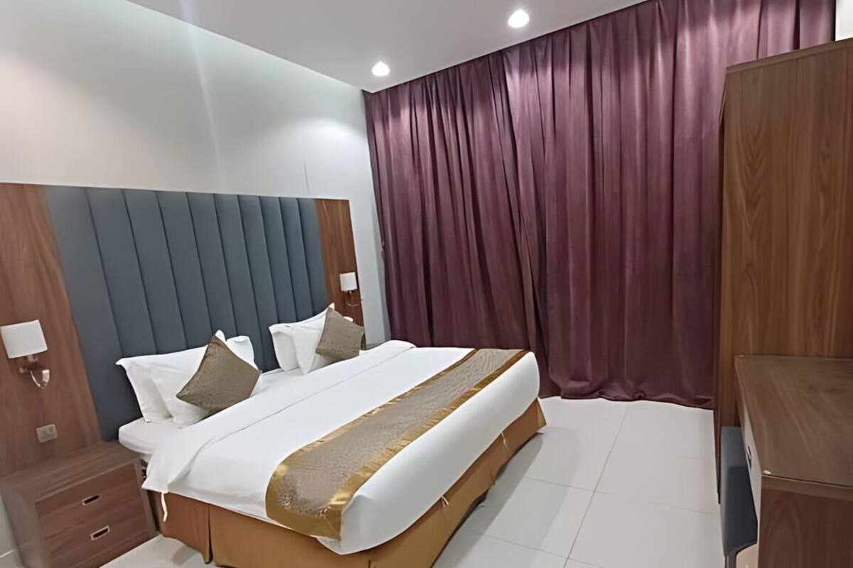 A modern hotel room with a large bed, wooden headboard, a wooden nightstand, a desk, and a large window with curtains.
