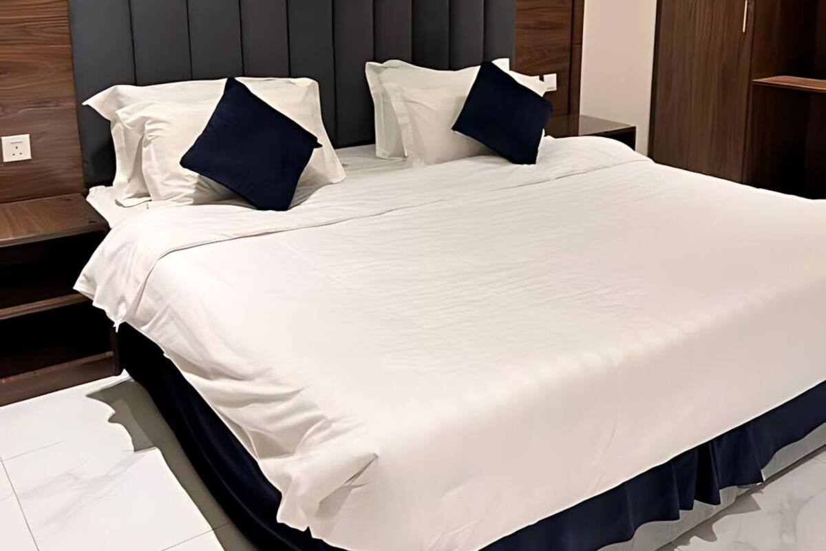 A neatly made bed with white and navy bedding, a dark headboard, and a wooden wardrobe in the background.