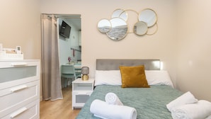 Ein ordentlich bezogenes Bett mit einer grünen Decke, einem braunen Kopfkissen und zwei aufgerollten Handtüchern. Ein weißer Nachttisch steht daneben, darüber an der Wand eine Lampe und ein Spiegel.