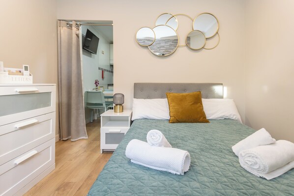 A neatly made bed with a green quilt, a brown pillow, and two rolled towels. There is a white nightstand with a lamp and a mirror on the wall above the bed.