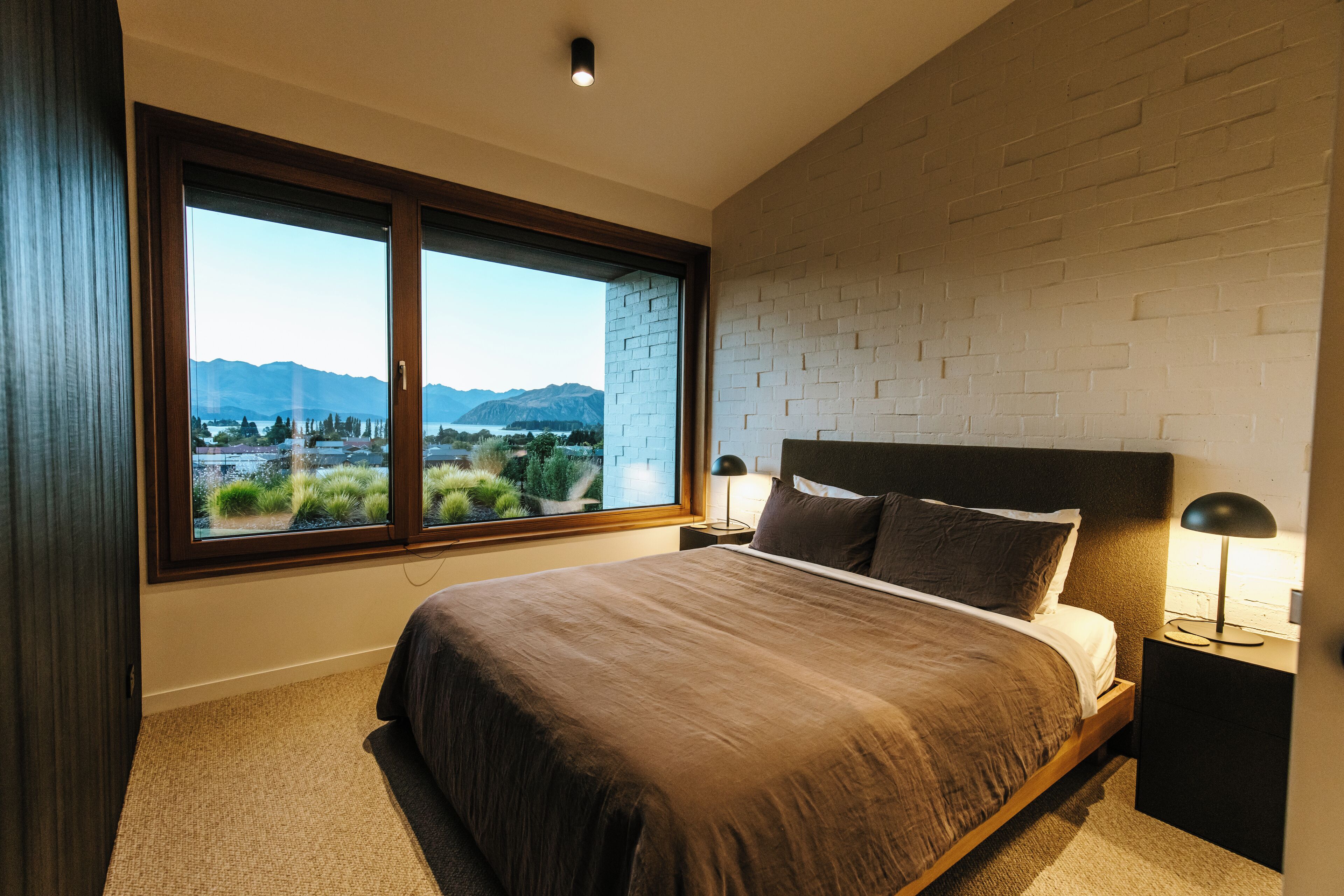 A bedroom with a large window, a bed, a nightstand, and a lamp.