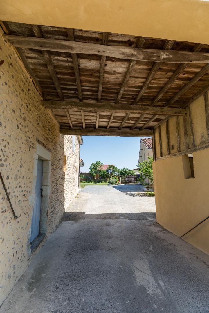 A narrow alleyway with a rustic stone wall on one side and a yellow wall on the other, leading to a paved area with buildings in the distance.