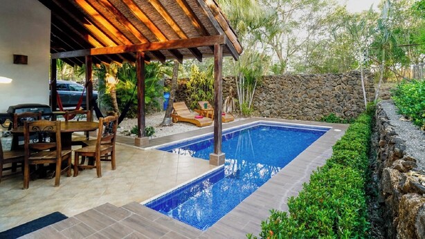 A swimming pool with a tiled deck, a covered seating area, and a stone wall in the background. - Casa Tropical - Fully equipped house with private pool and BBQ (Villareal Tamarindo)