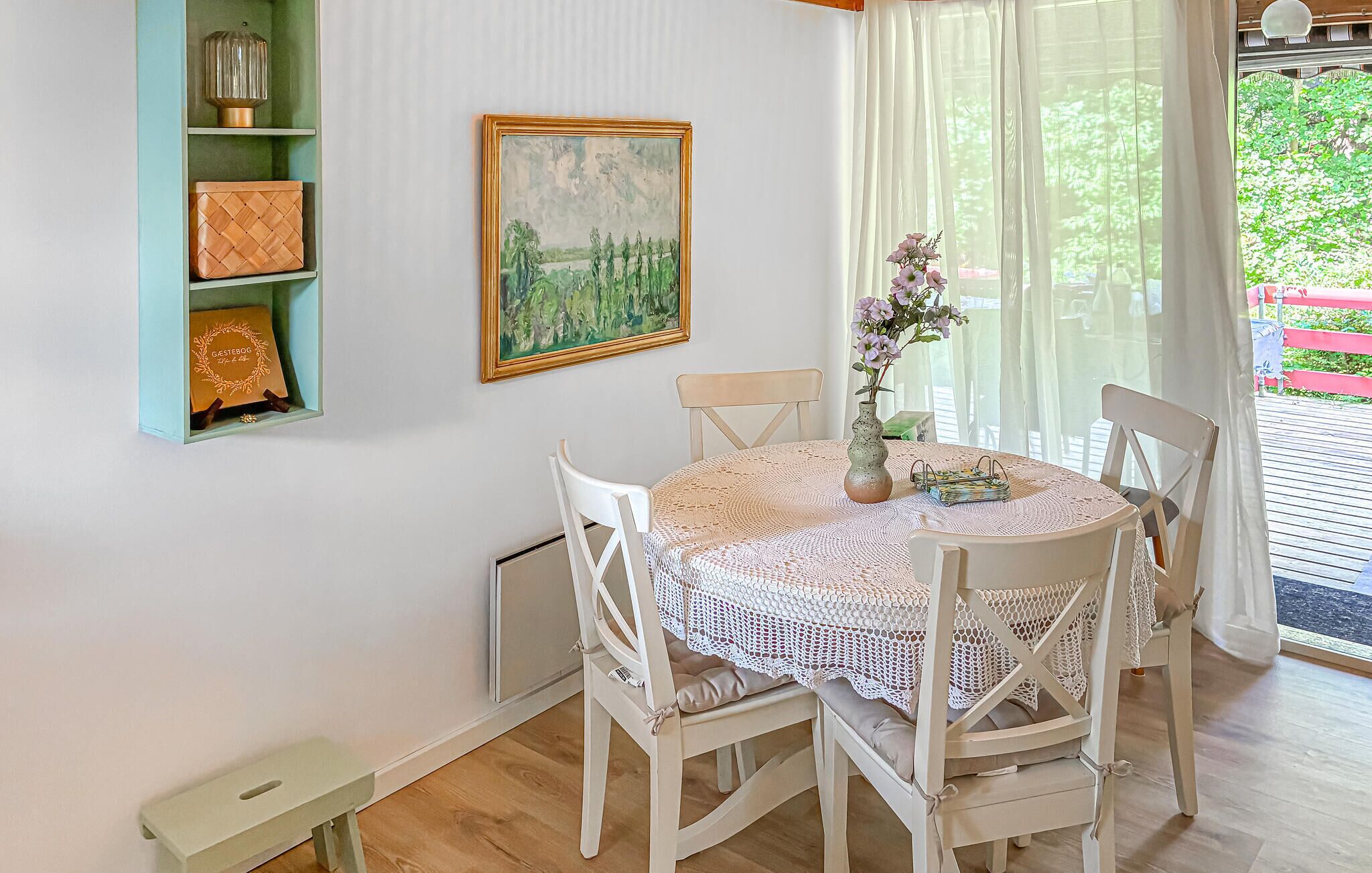 A dining area with a round table covered by a lace tablecloth, surrounded by white chairs. A painting hangs on the wall above the table, and a vase with flowers is on the table.