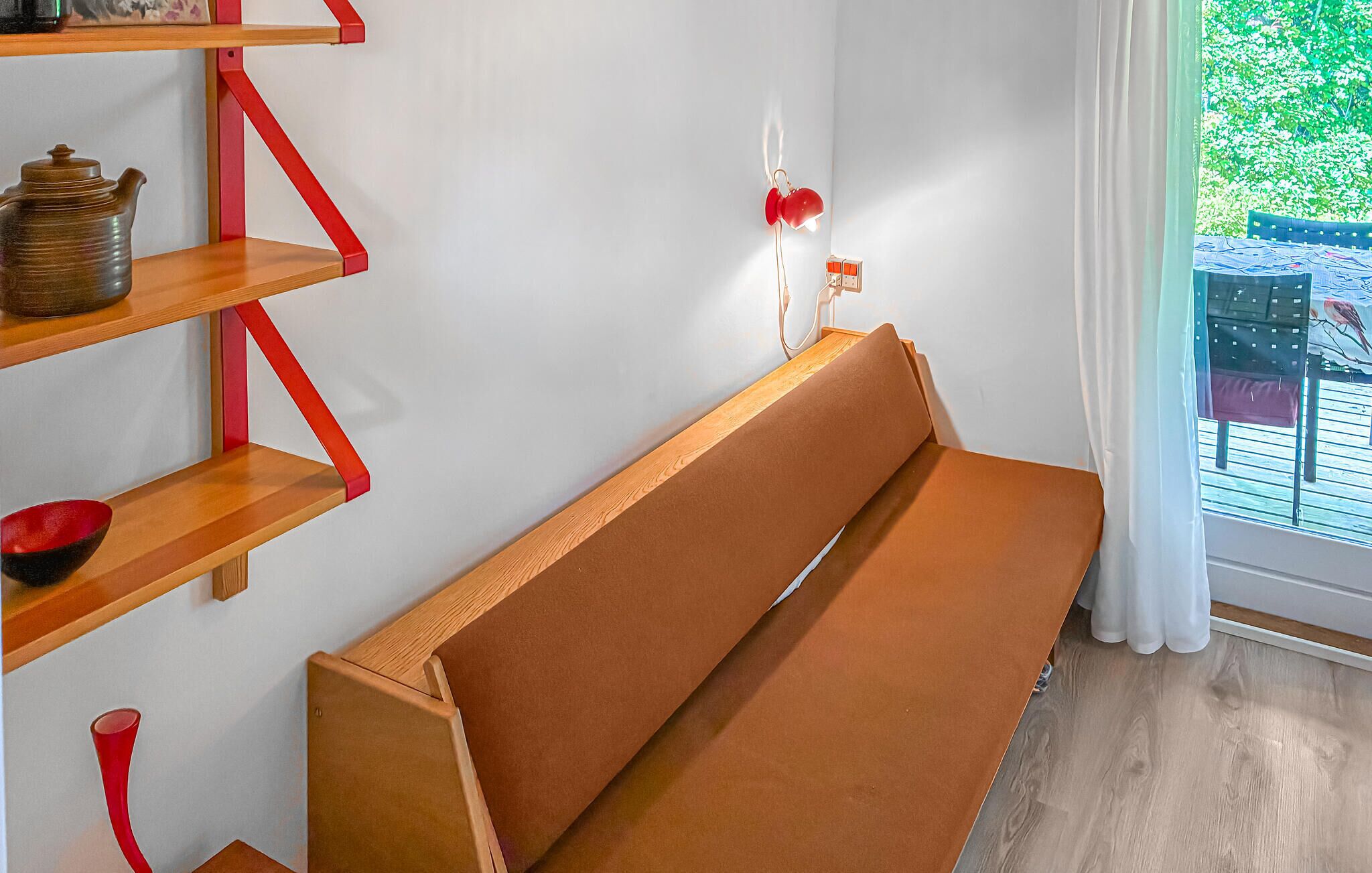 Una habitación con un banco de madera, estantes con detalles rojos y una ventana que da a zonas verdes.