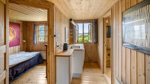 Une cabane en bois comprenant un lit, un coin cuisine et une fenêtre donnant sur un paysage verdoyant.