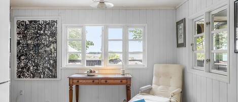 Una habitación con suelo de madera, una silla blanca, una mesa con lámpara y un ventanal con vistas a árboles y edificios.