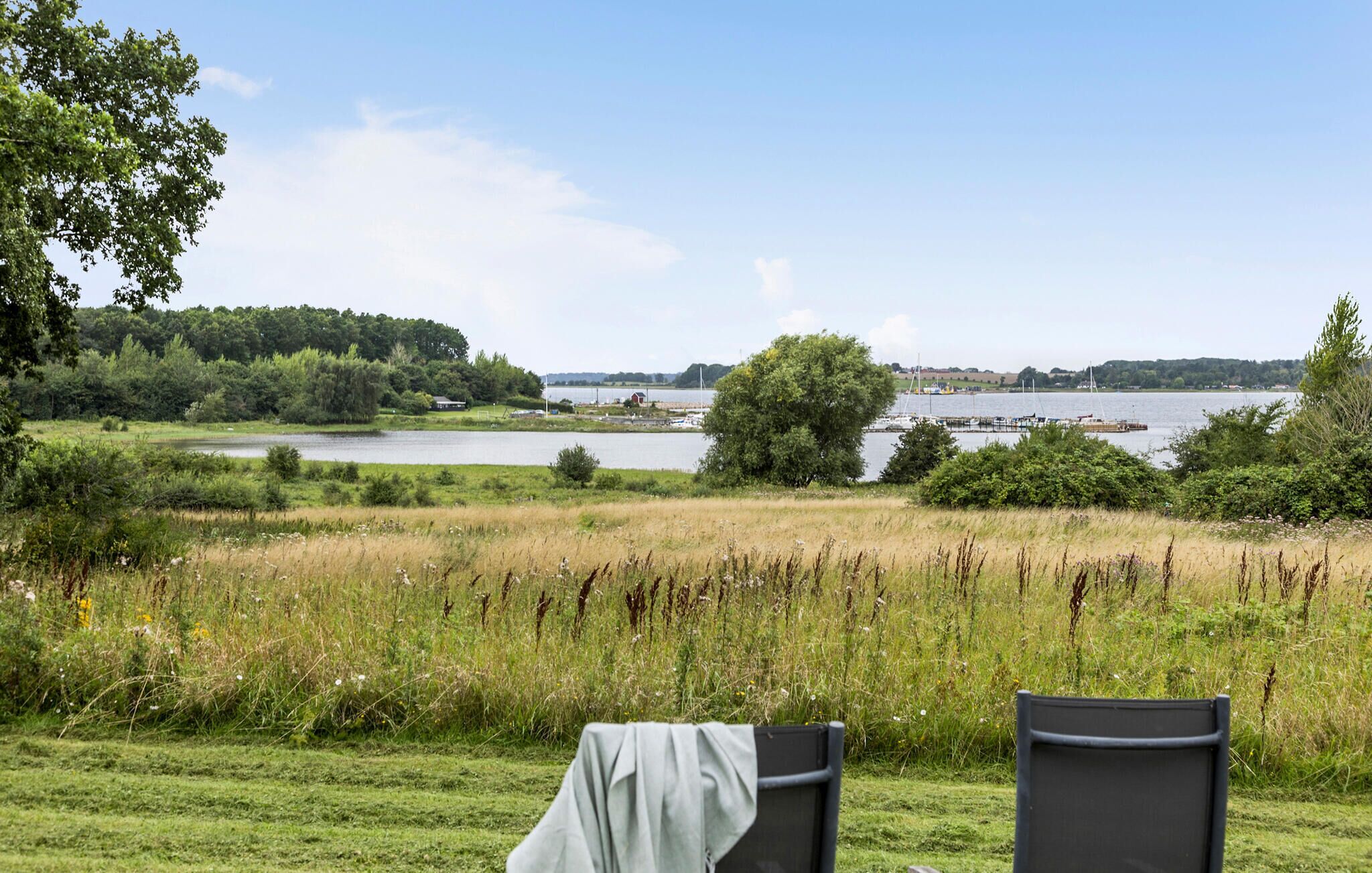 A lakeside view with two chairs and a table, surrounded by greenery and a clear sky.