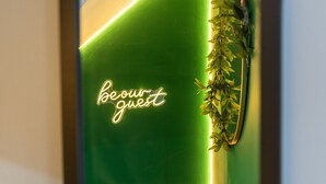 A room with a green wall, a neon sign reading "be our guest," and a plant decoration.