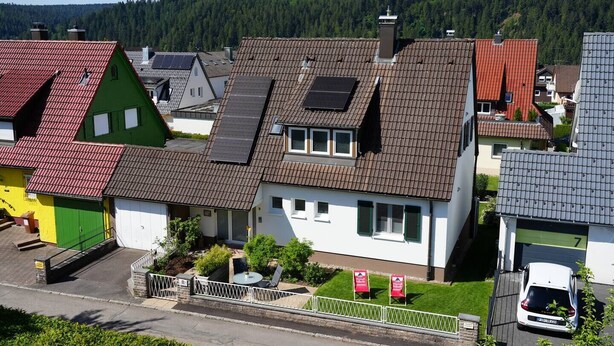 Et boligområde med huse med skrå tage og solpaneler, omgivet af grønt og en skovklædt bakke.