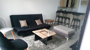 Ein Wohnzimmer mit einer schwarzen Couch, einem kleinen Holztisch, einem weißen Sessel und einem grauen Pouf. Es gibt eine Küchenzeile mit einem Barhocker und einem runden Holztisch.