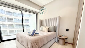 Une chambre moderne avec un grand lit, une table de chevet, une fenêtre donnant sur des immeubles et un luminaire au plafond.