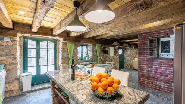 A rustic kitchen with a stone wall, wooden beams, a stone door, and a table with a basket of oranges.
