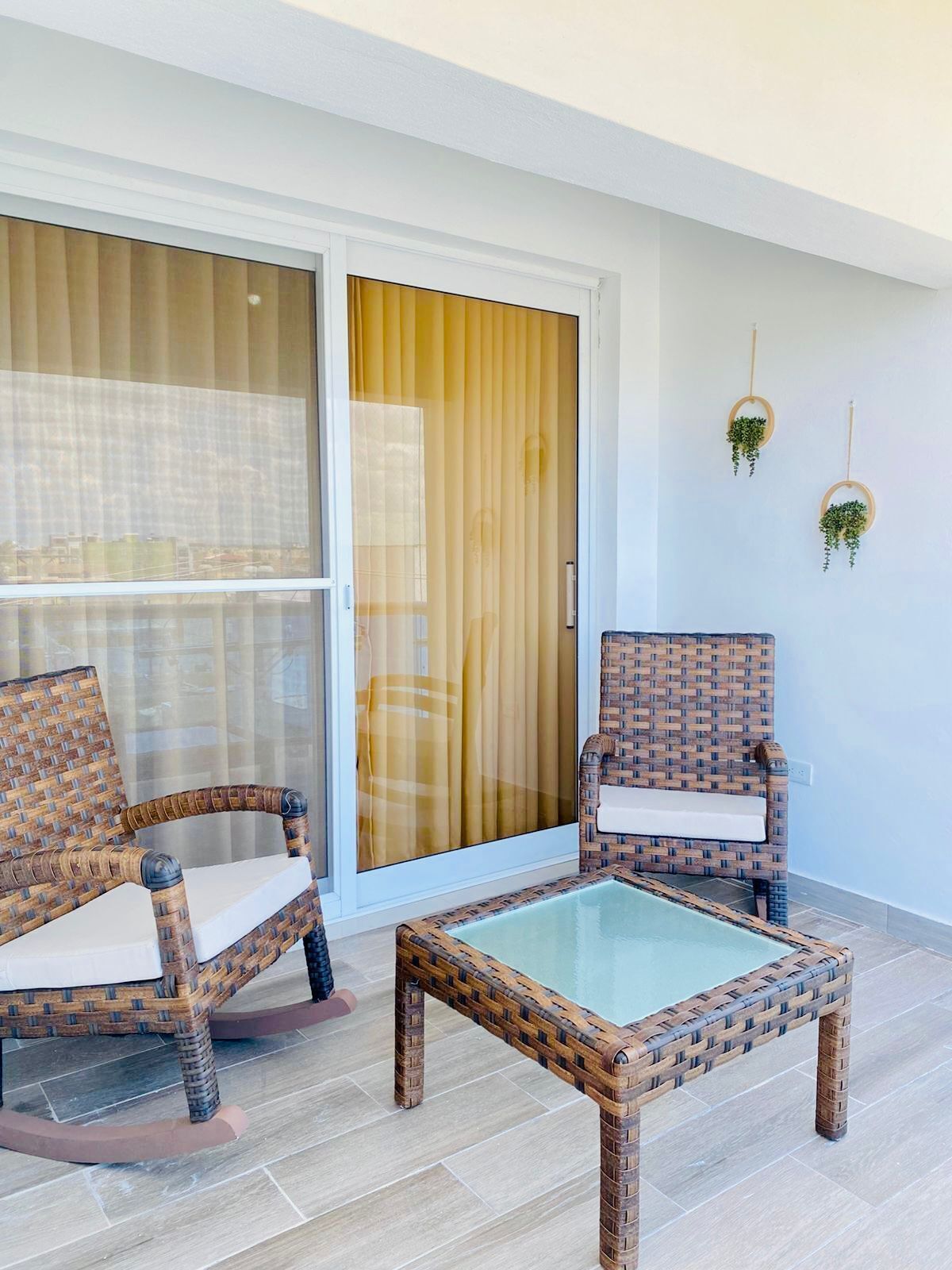Un balcon avec des fauteuils en osier, une table à plateau en verre et une porte-fenêtre coulissante avec des rideaux.