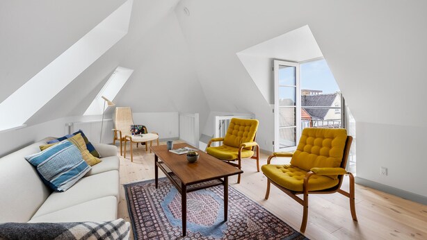 A spacious loft apartment with high ceilings, a wooden floor, a sofa, two yellow armchairs, a coffee table, and a large window offering a view of buildings.