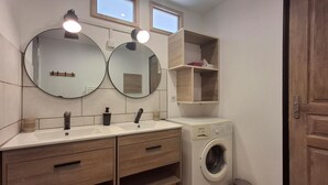 Une salle de bain avec deux lavabos, un miroir, un lave-linge et une petite poubelle.