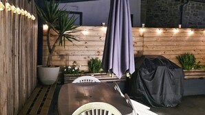 Une table de salle à manger en bois, des chaises, une plante en pot et une housse de protection pour barbecue.