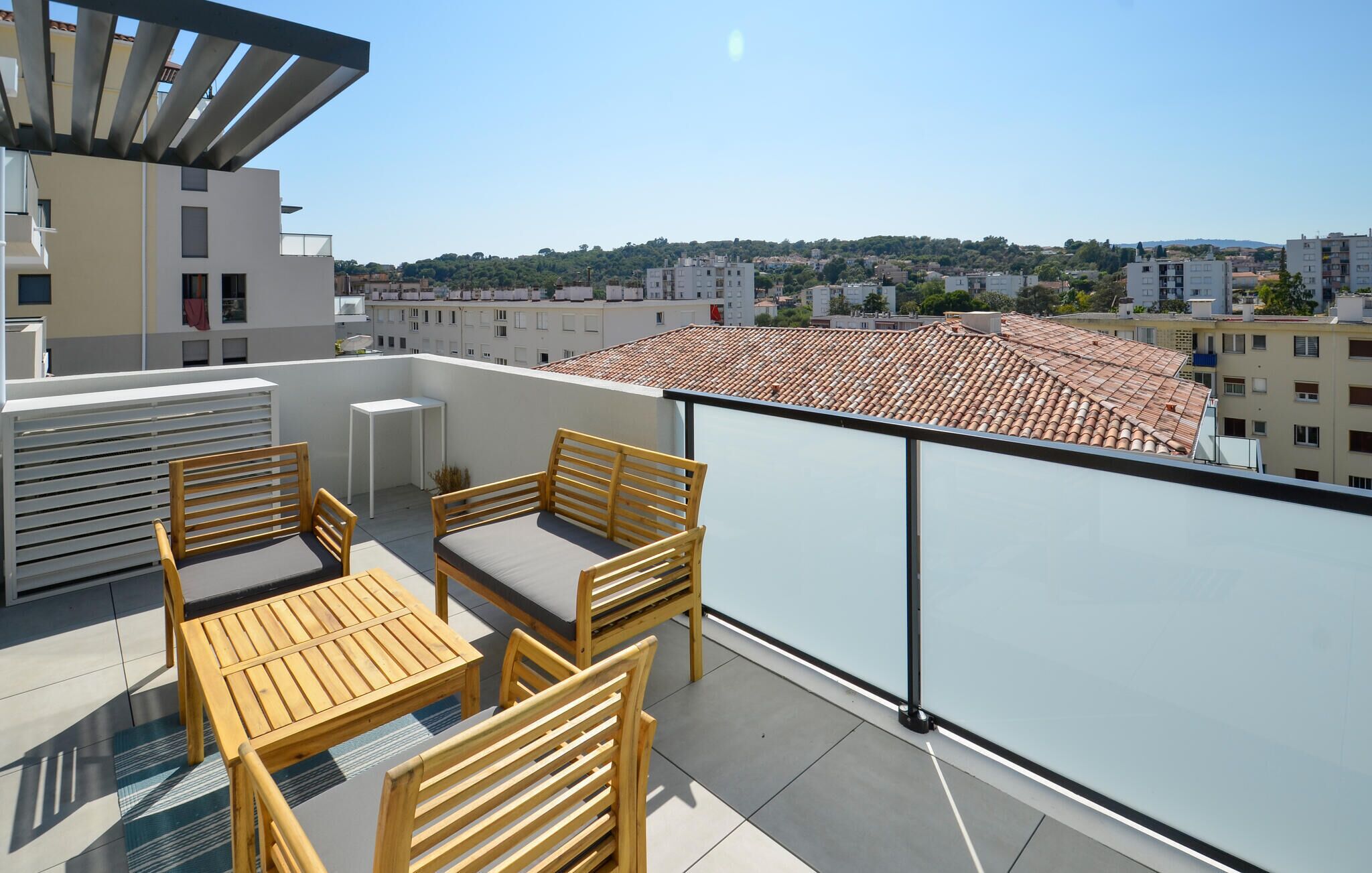 A rooftop terrace with wooden furniture, a glass balustrade, and a city view.