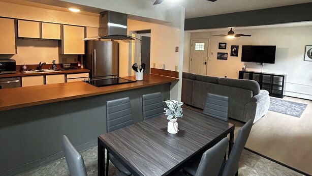 A modern kitchen with dark cabinetry, a central island, and a dining area with a table and chairs. - Beautiful Alaskan 2 Bedroom (Anchorage)