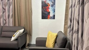 A modern living room with a grey sofa, a smaller armchair, and a yellow pillow. There is a painting on the wall and a window with curtains. - HideHaus Haven (Kumasi)