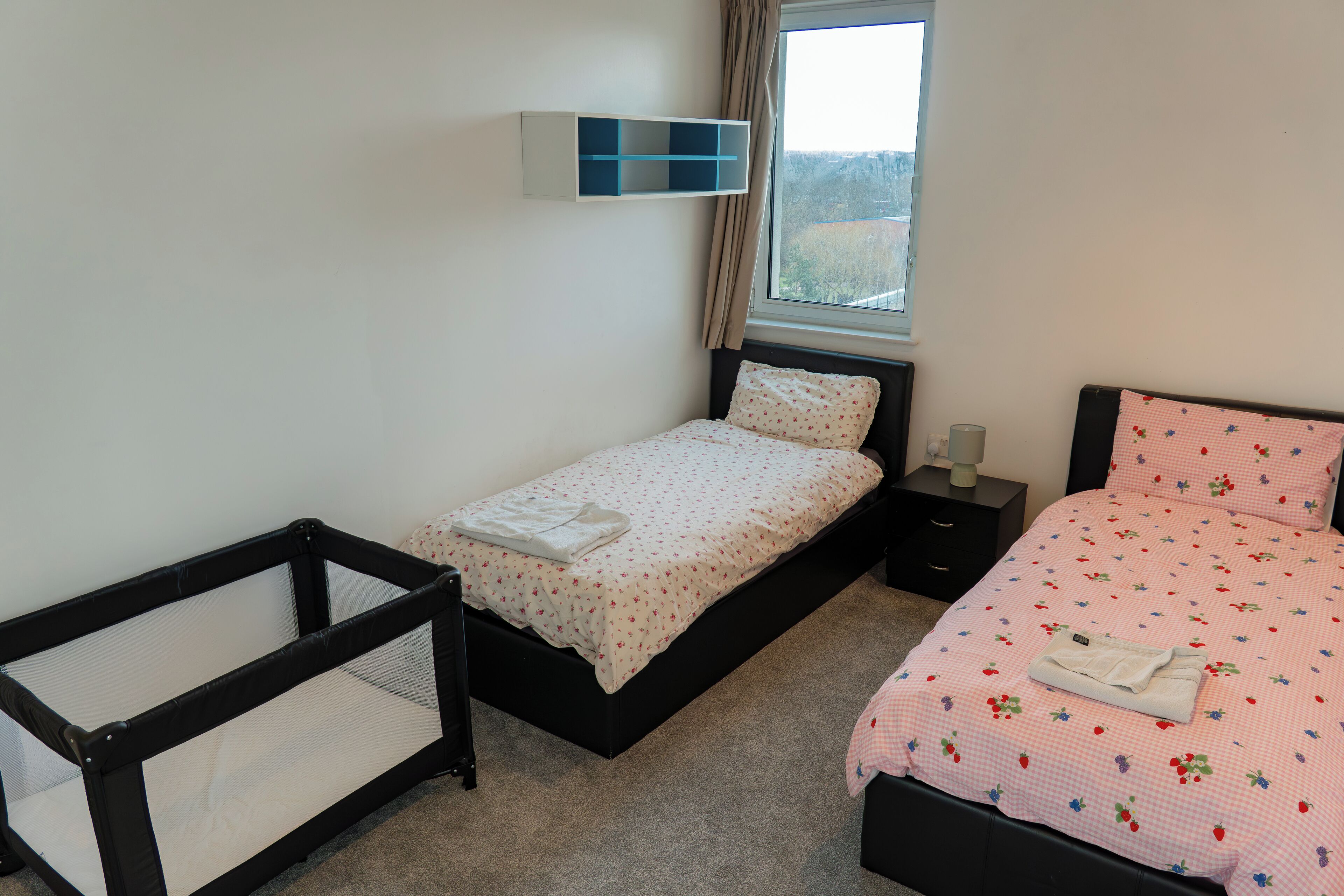 A bedroom with two beds, a window, a bedside table, and a wall-mounted shelf.