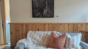 A grey sofa with a white fluffy blanket and two pillows, a framed picture of a mountain landscape on the wall, and a wooden floor.