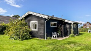 A dark wooden house with a sloped roof, a small porch with two white plastic chairs, and a well-maintained lawn. - Beautiful home in Otterup with WiFi (Otterup)