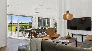 A modern living room with a sofa, a coffee table, and a TV. There is a dining area with chairs and a table visible through the glass doors. - Cozy home in Bogense with sauna (Bogense)