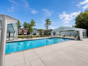 Une piscine entourée de parois en verre, un espace de détente couvert et des maisons en arrière-plan.