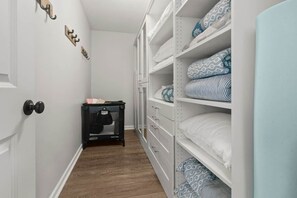 Un pasillo estrecho con paredes blancas, suelo de madera y un armario con estantes blancos y almohadones estampados.