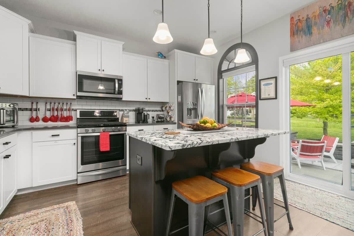 A modern kitchen with white cabinets, stainless steel appliances, a marble island with stools, and a large window offering a view of the outdoors.