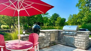 En udendørs terrasse med en sten-grill, et rundt bord med røde stole og en rød parasol.