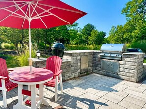 Eine Terrasse im Freien mit einem Stein-Barbecue, einem runden Tisch und roten Stühlen sowie einem roten Sonnenschirm.