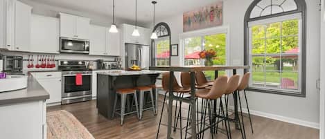 Una cocina moderna con armarios blancos, electrodomésticos de acero inoxidable, una isla central con taburetes y ventanas amplias con vistas a la vegetación.