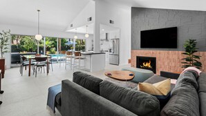A modern living room with a fireplace, a TV mounted on the wall, and a dining area with a pool view.