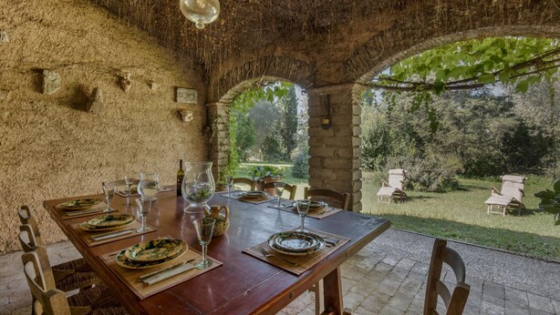 An outdoor dining area with rustic stone walls, a wooden table set for a meal, and a view of a lush garden.