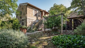 A rustic stone building with a wooden staircase leading to a door, surrounded by lush greenery and a stone pathway.