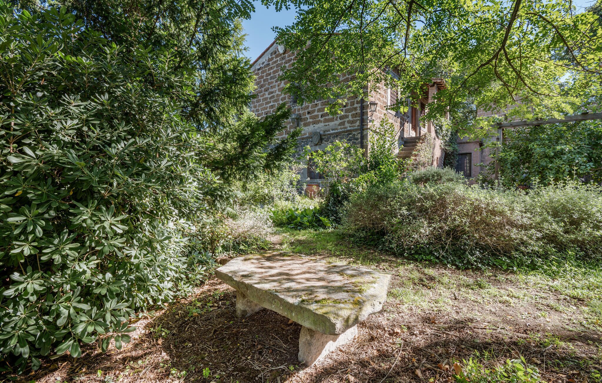 A stone bench in a garden with dense foliage and a historic building in the background.